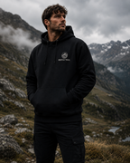 A man wearing a black pullover hoodie with a white circular 'MORPHATRAIL' logo on the chest, standing in a mountainous outdoor setting.