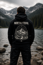 A man stands with his back to the camera wearing a black hoodie with a large white graphic print on the back that reads 'NORTH//TRAIL' and 'EXPLORING WEAVER MOUNTAIN'. He is positioned on a rocky shore in front of a lake and forested mountains.
