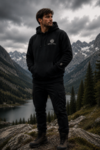 A man wearing a black pullover hoodie and black cargo pants stands on a rocky mountainside overlooking a lake and pine-covered valley under a cloudy sky. A small white chest logo is visible on the hoodie.