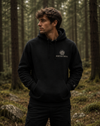 A man wearing a black pullover hoodie with a white circular Northway Trail logo on the left chest, standing in a dense forest.